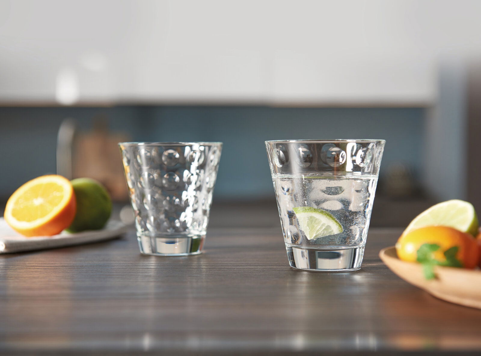 Zwei Trinkgläser Optic 215ml auf einem Tisch, eines gefüllt mit Wasser und Limettenscheibe, im Hintergrund unscharf Obst und Küche, seitliche Perspektive.