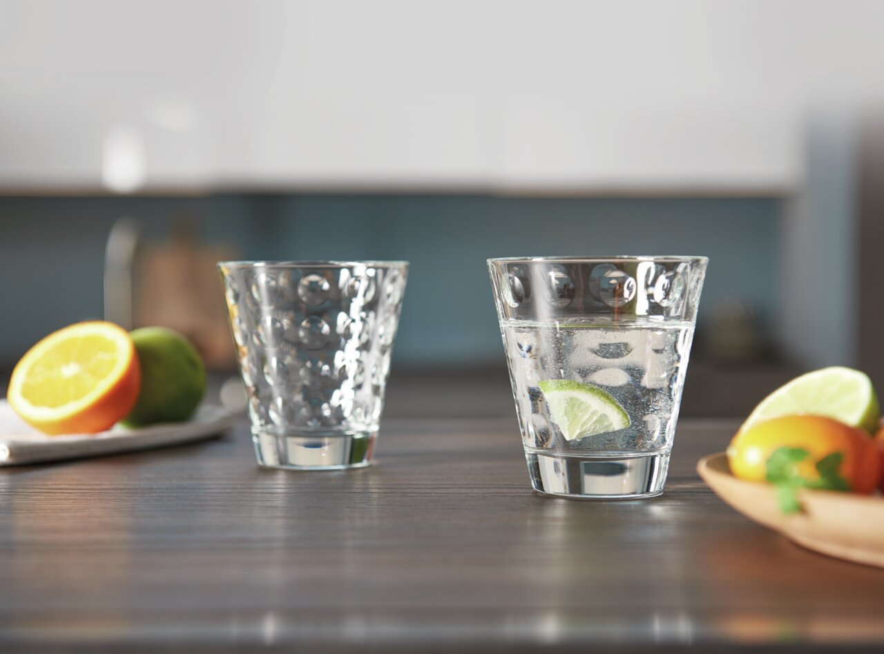 Zwei Trinkgläser Optic 215ml auf einem Tisch, eines gefüllt mit Wasser und Limettenscheibe, im Hintergrund unscharf Obst und Küche, seitliche Perspektive.