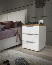 Moderne Nachtkommode mit drei Schubladen in Hochglanz-Weiß, seitliche Perspektive neben einem Bett, dekoriert mit Büchern und einer Lampe.