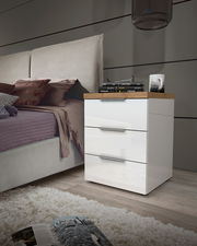 Moderne Nachtkommode mit drei Schubladen in Hochglanz-Weiß, seitliche Perspektive neben einem Bett, dekoriert mit Büchern und einer Lampe.