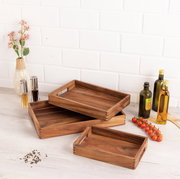 Drei hölzerne Tabletts in verschiedenen Größen, gestapelt auf einem hellen Holztisch, umgeben von Gewürzflaschen, Olivenölflaschen und einer Vase mit Blumen. Perspektive von oben.