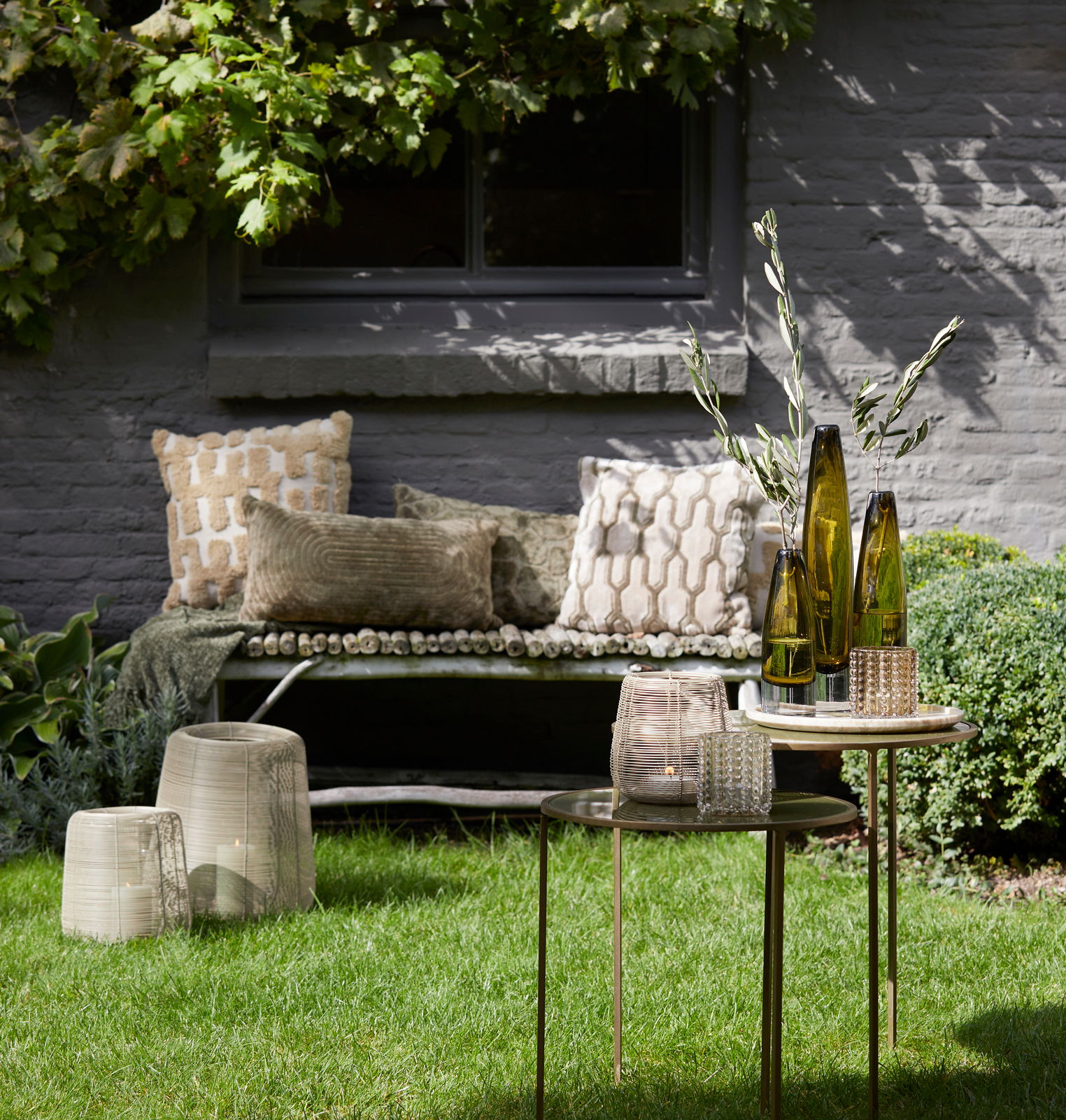 Light&Living Windlicht ADETA Gartenansicht mit dekorativen Windlichtern auf einem Tisch, umgeben von grüner Vegetation und gemütlichen Kissen auf einer Bank im Hintergrund.