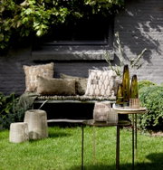 Gartenansicht mit dekorativen Windlichtern auf einem Tisch, umgeben von grüner Vegetation und gemütlichen Kissen auf einer Bank im Hintergrund.