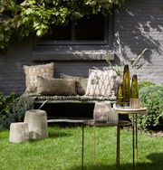 Gartenansicht mit dekorativen Windlichtern auf einem Tisch, umgeben von grüner Vegetation und gemütlichen Kissen auf einer Bank im Hintergrund.