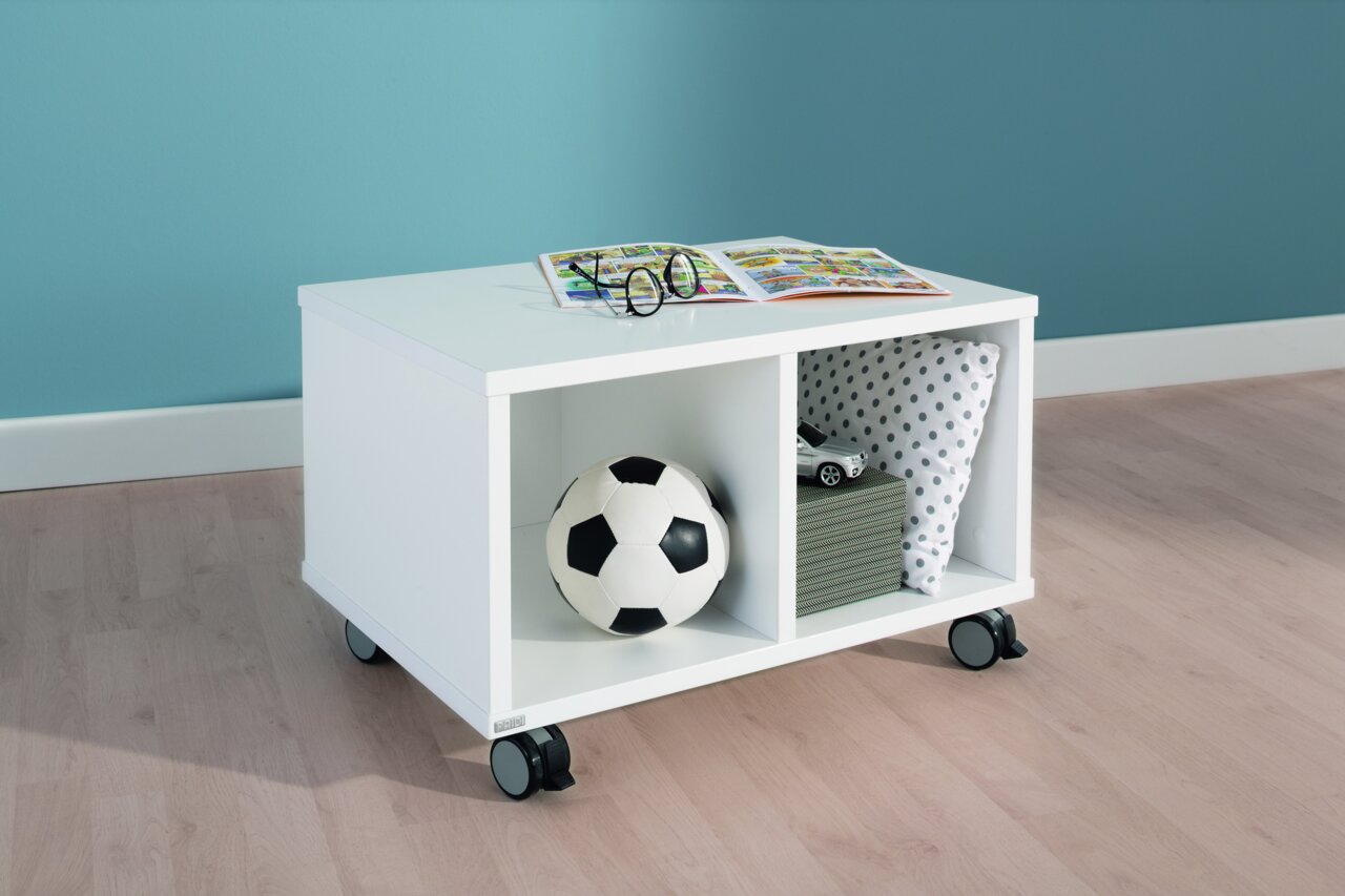 Weißes Rollregal mit zwei Fächern, gefüllt mit einem Fußball, Büchern und einem Kissen, auf Rollen, seitliche Perspektive.