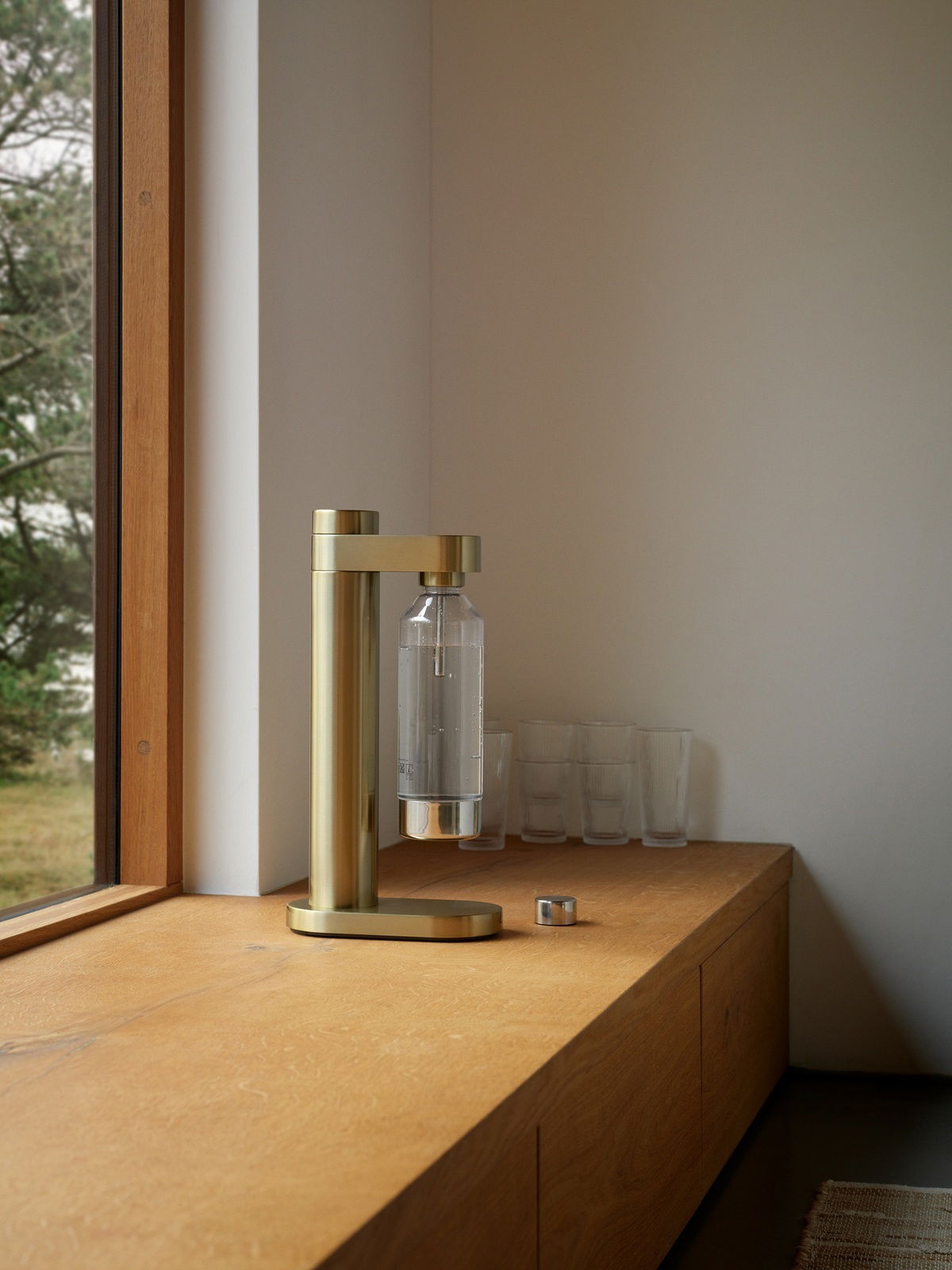 Stelton Wassersprudler BRUS Goldener Wassersprudler auf einer Holzoberfläche, seitliche Perspektive, mit einem Fenster im Hintergrund.