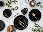 Draufsicht auf ein elegantes Tisch-Arrangement mit schwarzen Tellern und Bechern, goldfarbenem Besteck, Kerzen und Gläsern auf hellem Untergrund, dekoriert mit grünen Zweigen