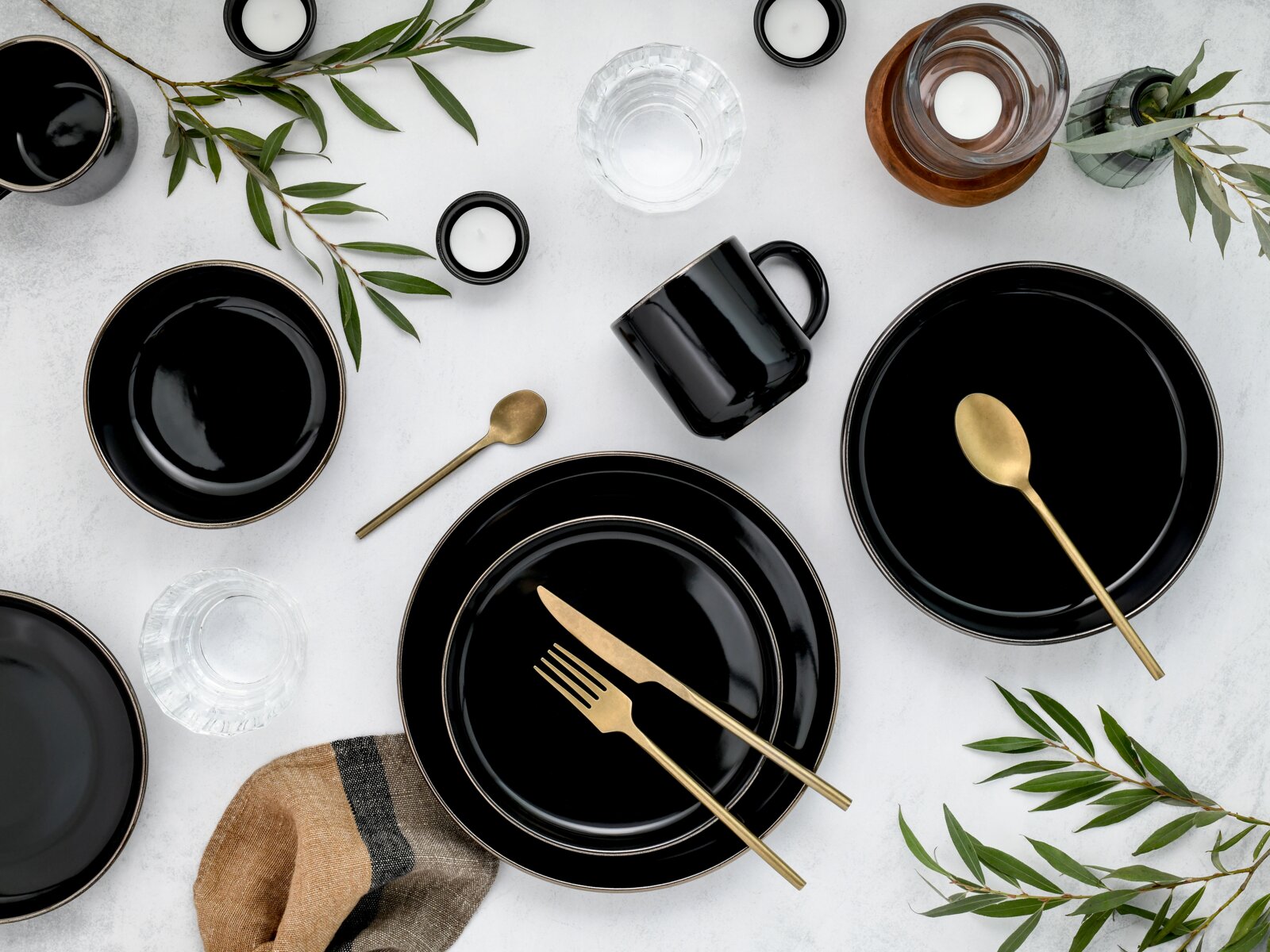 Draufsicht auf ein elegantes Tisch-Arrangement mit schwarzen Tellern und Bechern, goldfarbenem Besteck, Kerzen und Gläsern auf hellem Untergrund, dekoriert mit grünen Zweigen