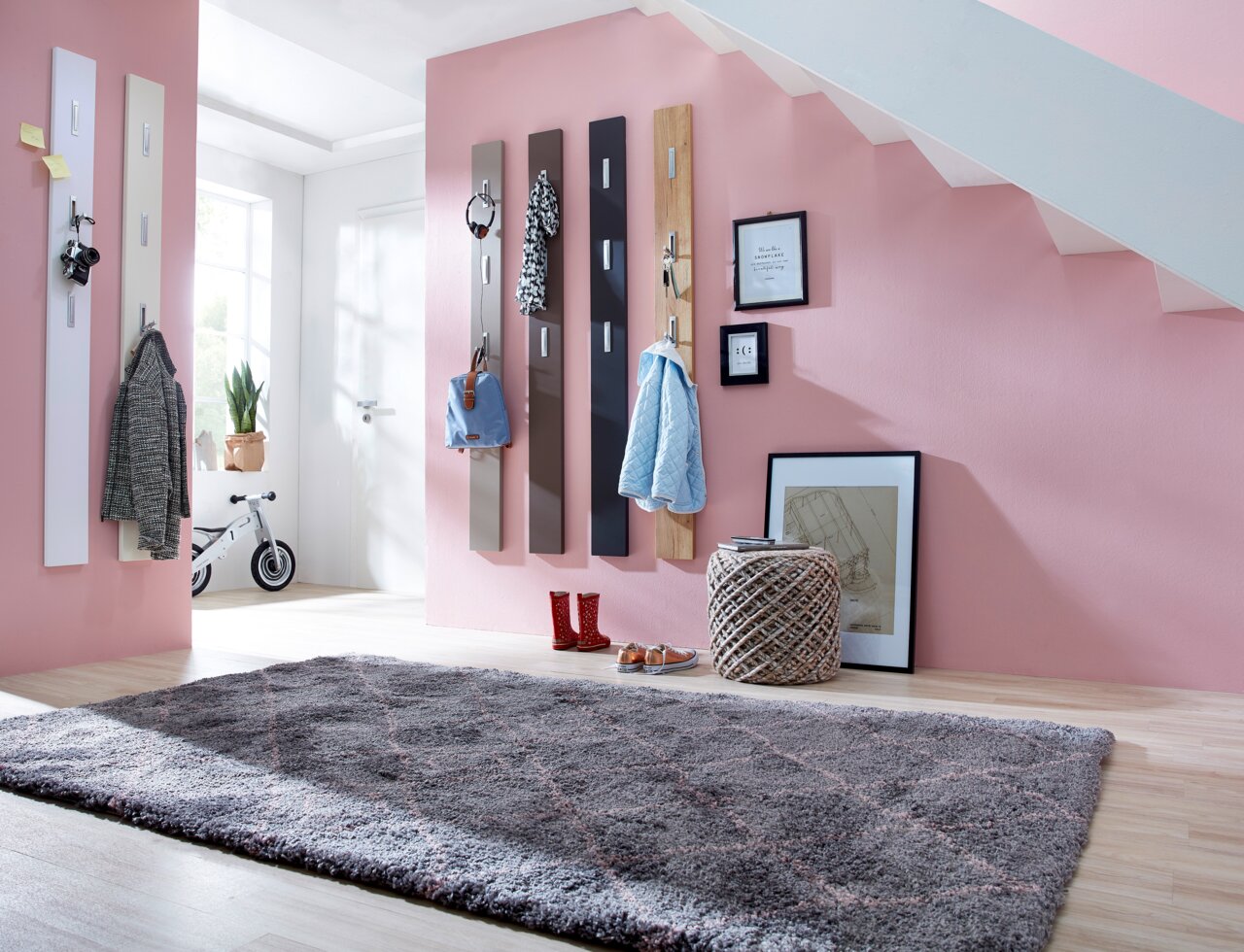 XORA Garderobepaneel AVILA Moderne garderobepanelen in verschillende kleuren tegen een roze muur, volgehangen met jassen en accessoires, in een lichte gang met een trap op de achtergrond, gezien vanuit een zijdelingse hoek.