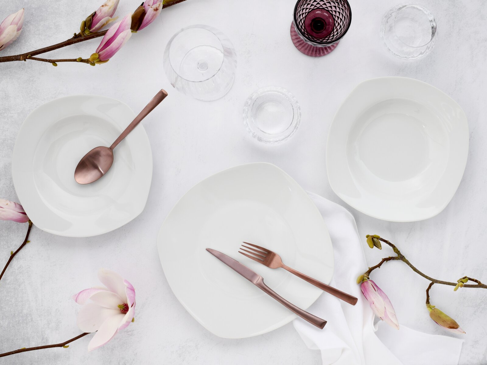 Draufsicht (Flatlay) auf weißes, quadratisch abgerundetes Porzellan-Geschirrset mit Teller und Schalen, roségoldenem Besteck, Glas und Magnolienzweigen auf hellem Hintergrund.