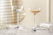 Zwei elegante Likörschalen aus der Serie POESIA mit 90 ml Fassungsvermögen auf einem Marmortisch, eine gefüllt mit Likör, im Hintergrund unscharfe goldene Akzente und weiße Blumen, seitliche Perspektive.