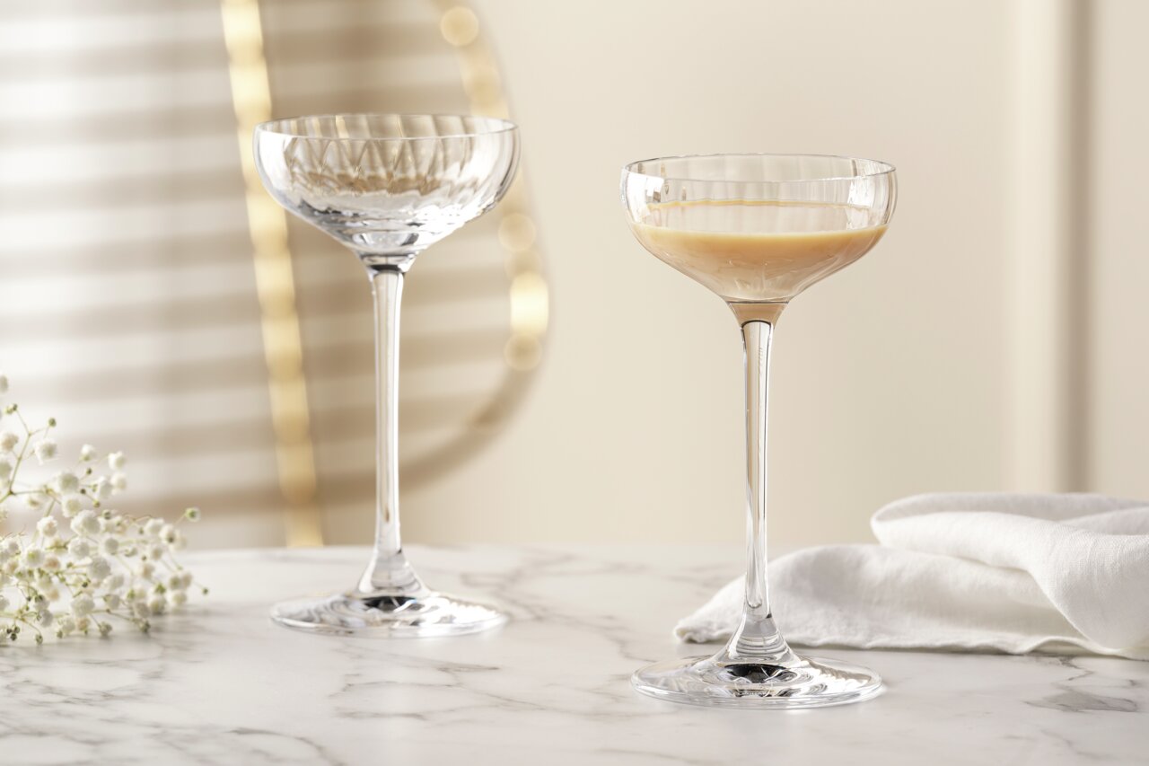 Zwei elegante Likörschalen aus der Serie POESIA mit 90 ml Fassungsvermögen auf einem Marmortisch, eine gefüllt mit Likör, im Hintergrund unscharfe goldene Akzente und weiße Blumen, seitliche Perspektive.