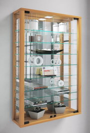 Hohe Glasvitrine mit hellem Holzrahmen, Glasböden und LED-Beleuchtung, gefüllt mit Deko-Objekten und Büchern, in schräger Frontansicht von links.