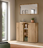 Frontale Ansicht eines modernen Holz-Sideboards mit vertikaler Lamellenoptik, zwei Türen und offenem Mittelregal, darüber ein rechteckiger Wandspiegel im Holzrahmen in einem hellen Wohnraum.