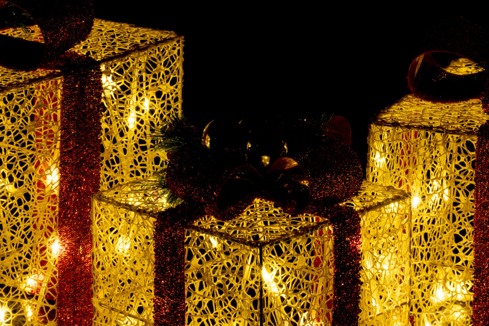 Leuchtende, goldene Deko-Geschenkboxen mit warmweißen Lichtern und roten Glitzerbändern vor schwarzem Hintergrund, Perspektive: leicht schräg von vorne.