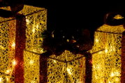 Leuchtende, goldene Deko-Geschenkboxen mit warmweißen Lichtern und roten Glitzerbändern vor schwarzem Hintergrund, Perspektive: leicht schräg von vorne.