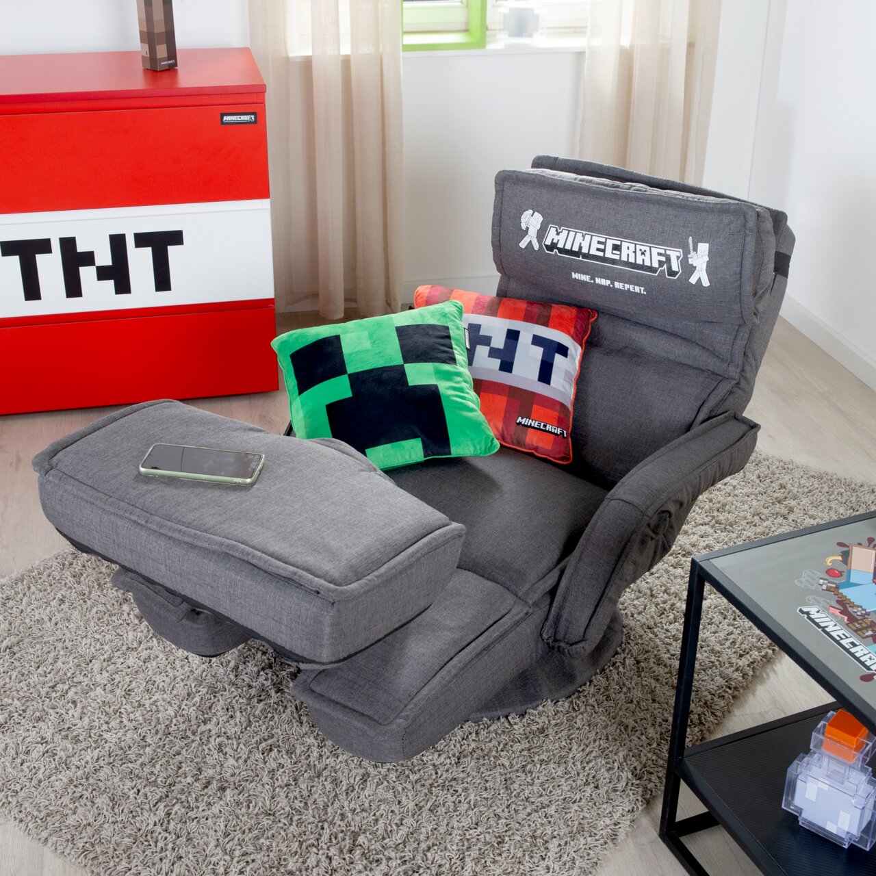 Grauer Minecraft-Gaming-Sessel mit verstellbarer Rückenlehne und ausgeklappter Fußstütze, mit Creeper- und TNT-Kissen, in schräger Frontansicht im Wohnzimmer auf Teppich
