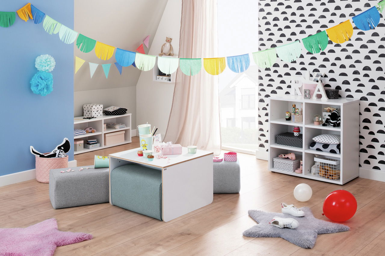 Kinderzimmer mit einem weißen Kindertisch und grauen Sitzpolstern, dekoriert mit bunten Girlanden und Spielzeug, aus der Perspektive eines Erwachsenen betrachtet.