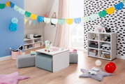 Kinderzimmer mit einem weißen Kindertisch und grauen Sitzpolstern, dekoriert mit bunten Girlanden und Spielzeug, aus der Perspektive eines Erwachsenen betrachtet.