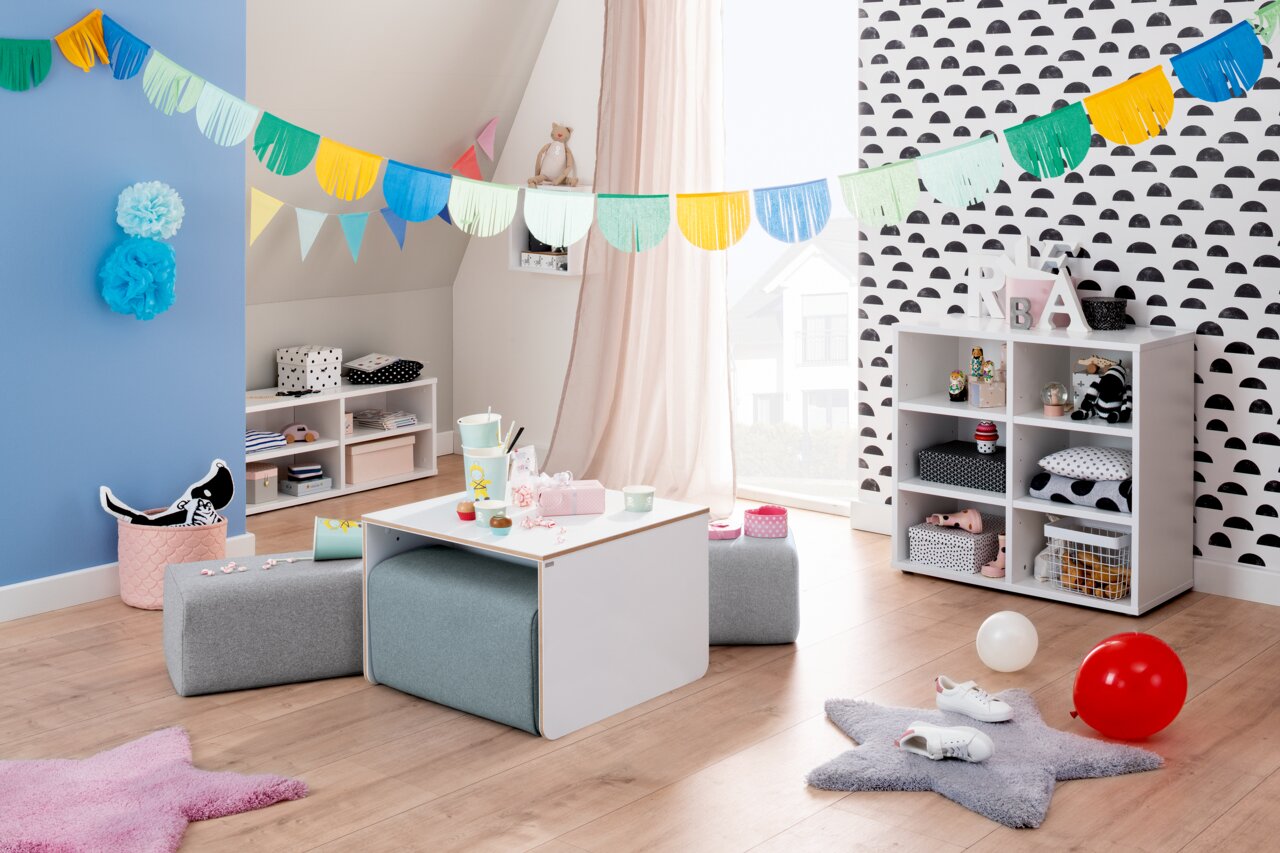 Kinderzimmer mit einem weißen Kindertisch und grauen Sitzpolstern, dekoriert mit bunten Girlanden und Spielzeug, aus der Perspektive eines Erwachsenen betrachtet.