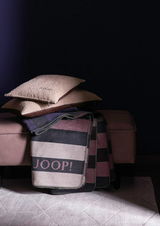 Zwei beige Zierkissen mit JOOP!-Logo auf einem Sofa, darüber eine gestreifte Decke mit JOOP!-Schriftzug, seitliche Perspektive.