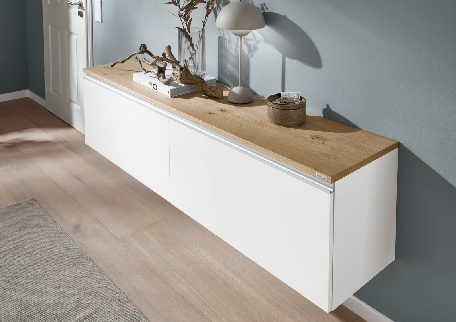 Modernes, wandhängendes Sideboard in Weiß mit Eichenholz-Oberplatte, dekoriert mit Lampe, Vase und Dekoobjekten, in schräger Frontansicht von rechts.
