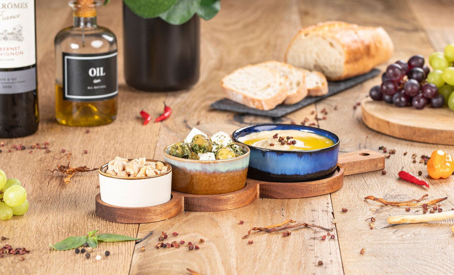 Schalen-Set für Antipasti auf Holztablett mit Nüssen, Oliven und Dip auf rustikalem Holztisch, umgeben von Brot, Trauben, Wein und Olivenöl, aufgenommen aus leicht erhöhter schräger Perspektive.