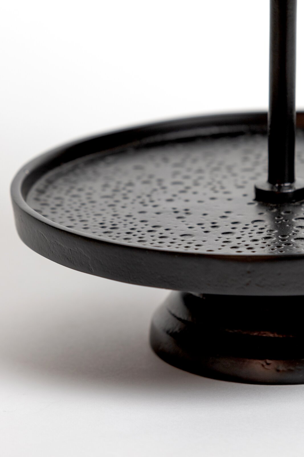 Nahaufnahme aus leicht seitlicher Perspektive eines schwarzen runden Metall-Beistelltisches mit strukturierter Oberfläche, erhöhtem Rand und stabilem Standfuß
