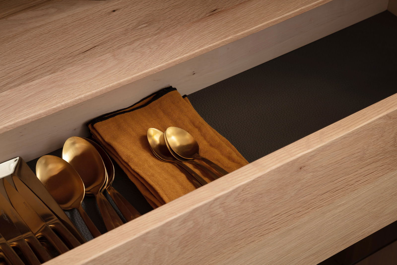 Nahaufnahme einer geöffneten Schublade aus Holz mit goldfarbenem Besteck und einem orangefarbenen Tuch, von oben betrachtet.