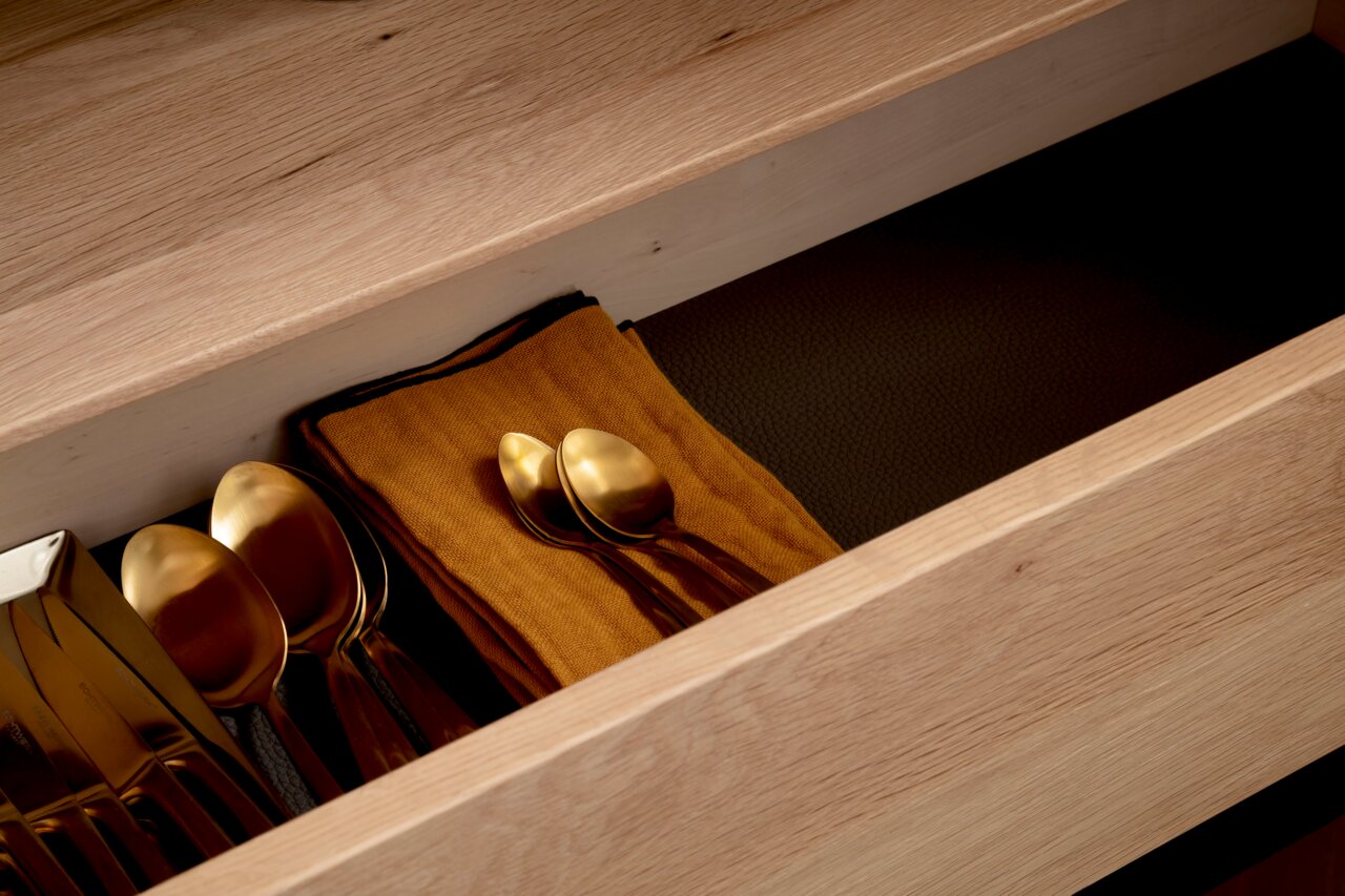 Nahaufnahme einer geöffneten Schublade aus Holz mit goldfarbenem Besteck und einem orangefarbenen Tuch, von oben betrachtet.