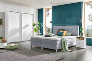 Modernes Boxbett in einem stilvollen Schlafzimmer mit grauem Polster, umgeben von Pflanzen und minimalistischen Möbeln, seitliche Perspektive.