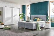 Modernes Boxbett in einem stilvollen Schlafzimmer mit grauem Polster, umgeben von Pflanzen und minimalistischen Möbeln, seitliche Perspektive.