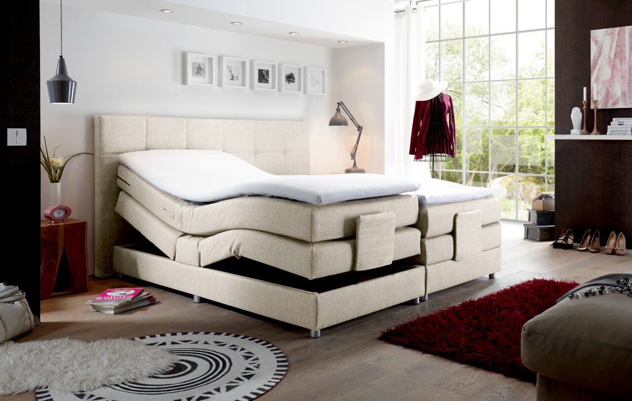 Boxspringbett in modernem Schlafzimmer, seitliche Perspektive, mit verstellbaren Matratzen und hellem Stoffbezug.