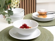 Weiße Porzellan-Schale auf passendem Teller mit roten Johannisbeeren und Müsli, daneben gestapelte weiße Schüsseln und Teller mit Gebäck auf grünen Platzsets, aufgenommen aus leicht schräger Frontperspektive.