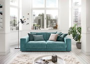 Zweiersofa in Türkis mit mehreren Kissen, in einem hellen Wohnzimmer mit großen Fenstern und moderner Dekoration, Frontalansicht.
