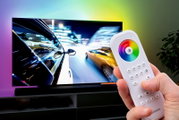 Close-up van een hand die een afstandsbediening met een kleurencirkel en knoppen vasthoudt, voor een televisie met een dynamisch beeld. Perspectief: van voren gericht op de afstandsbediening.