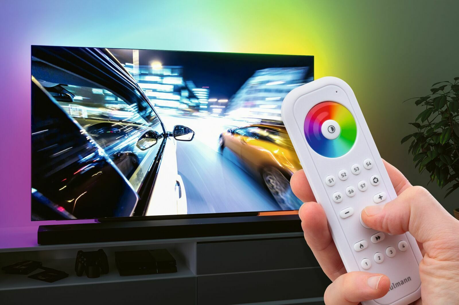 Nahaufnahme einer Hand, die eine Fernbedienung mit Farbkreis und Tasten hält, vor einem Fernseher mit dynamischem Bild. Perspektive: von vorne auf die Fernbedienung gerichtet.