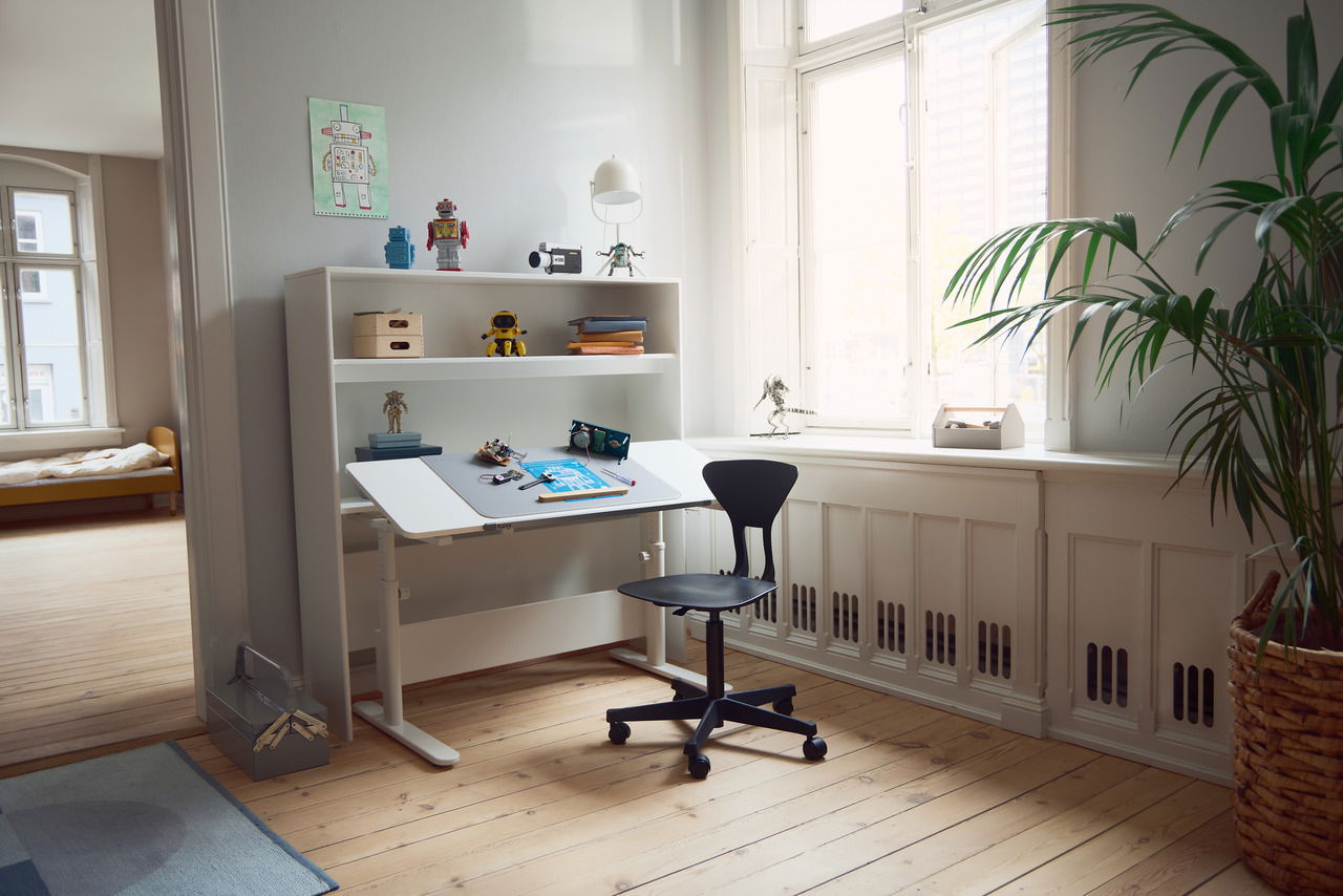 Moderner Schreibtisch mit verstellbarer Tischplatte, umgeben von Regalen mit Spielzeugrobotern, in einem hellen Raum mit Holzfußboden und großen Fenstern. Perspektive von der Seite.