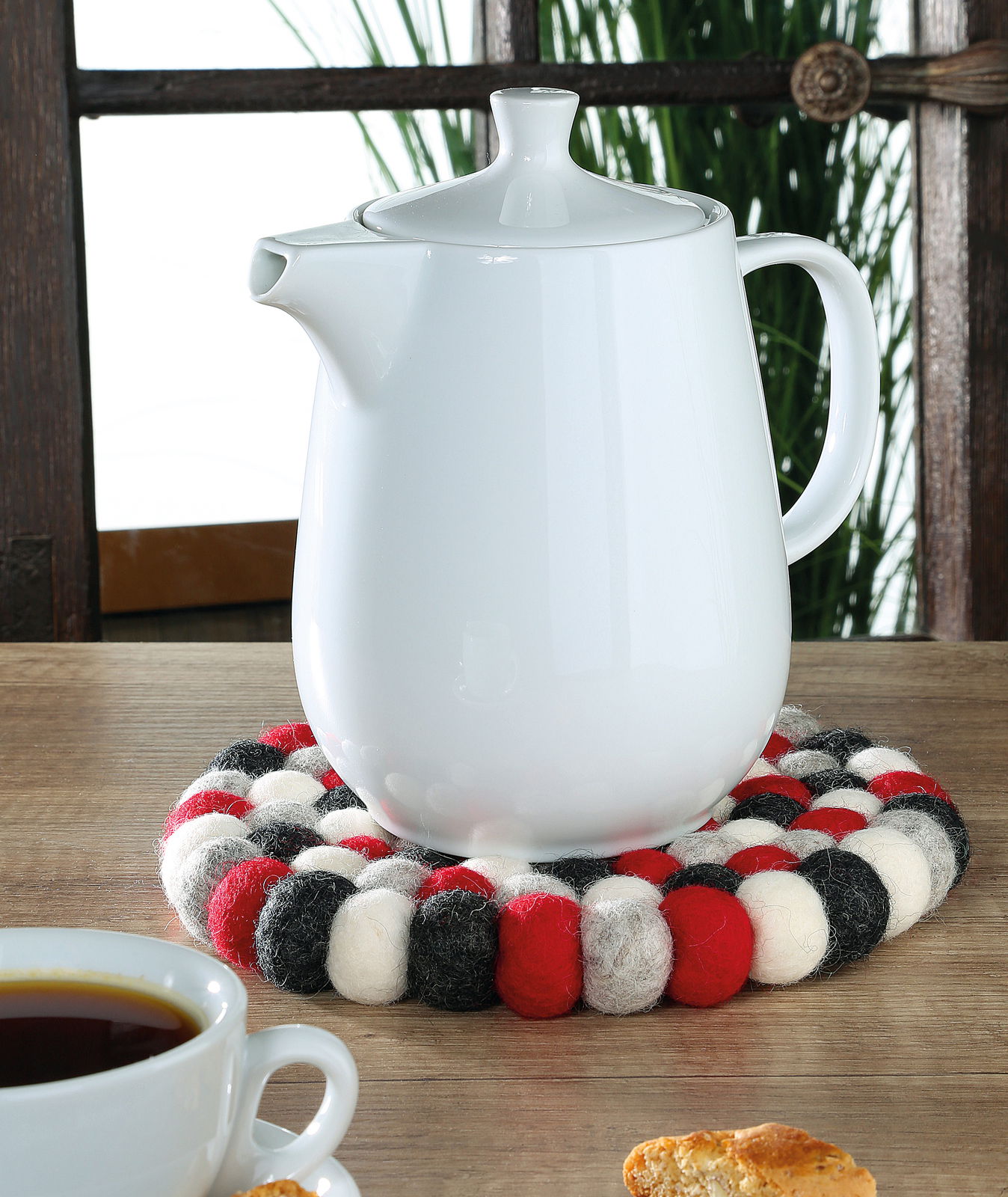 Runder Untersetzer LANA ROSSO aus Filzkugeln in Rot, Schwarz und Weiß, mit einer weißen Teekanne darauf, aus der Perspektive von vorne fotografiert.