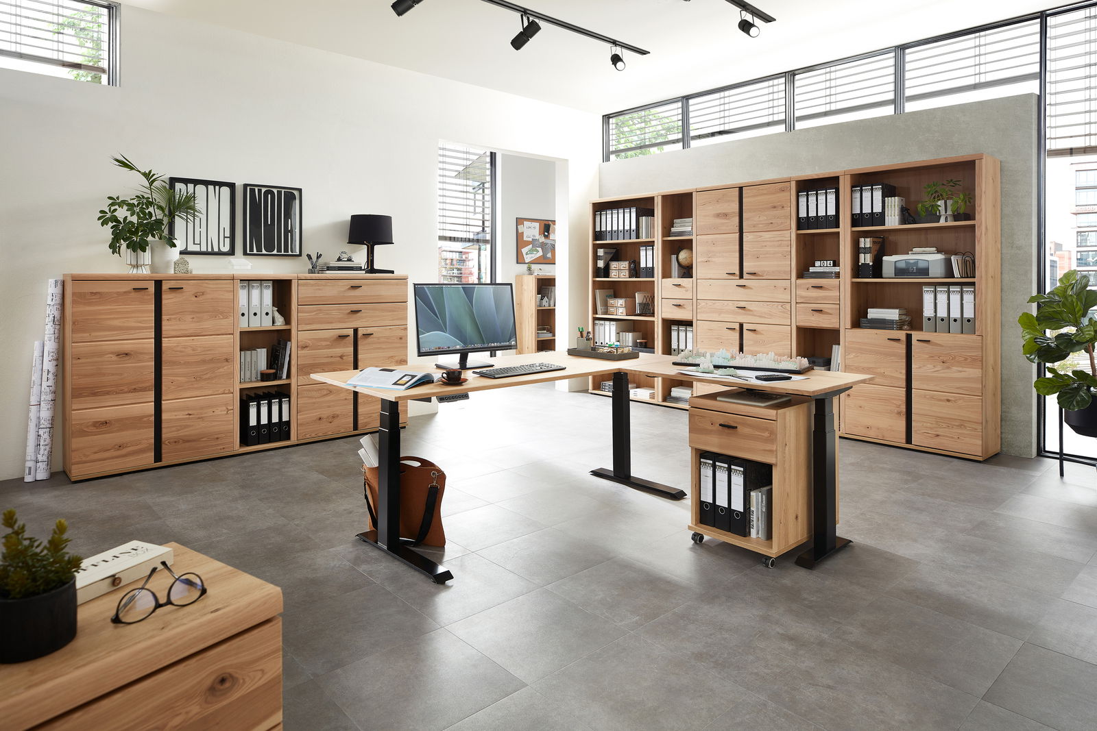 Moderne Büromöbel in einem hellen Raum mit großen Fenstern, bestehend aus einem höhenverstellbaren Schreibtisch, mehreren Aktenschränken und Regalen aus Holz. Perspektive: Frontalansicht.