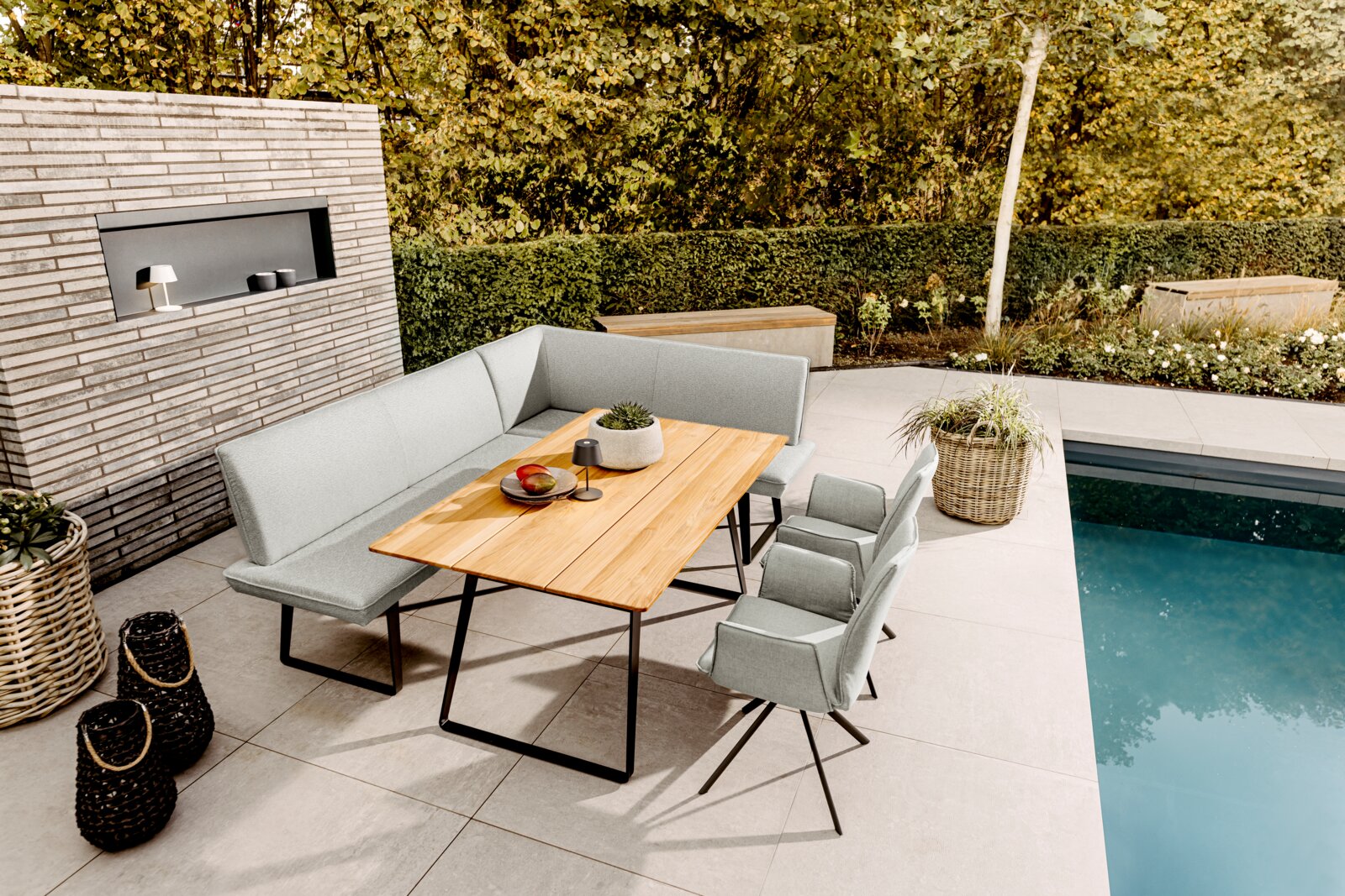 Modernes Outdoor-Ess-Set auf einer Poolterrasse: graue gepolsterte Eckbank mit Holztisch und zwei Stühlen, fotografiert aus leicht erhöhter schräger Perspektive von vorne links.