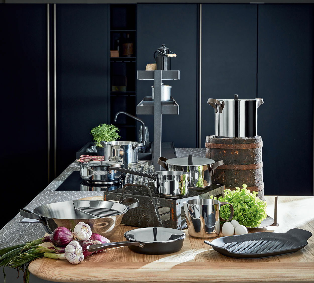 Küchenansicht mit einem 7-teiligen Topfset Edo aus Edelstahl, bestehend aus verschiedenen Töpfen und Pfannen, auf einer modernen Küchenarbeitsplatte arrangiert. Perspektive von vorne.