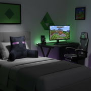 Modernes Gaming-Schlafzimmer mit Bett und Minecraft-Kissen im Vordergrund, Schreibtisch mit Monitor und RGB-Beleuchtung sowie Gaming-Stuhl rechts; Perspektive: leicht schräg von vorne aus dem Raum.