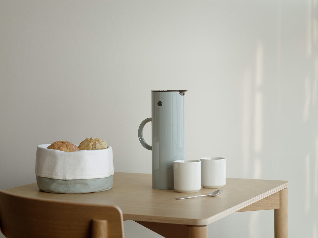 Stelton Isolierkanne 1 Liter Dusty Green Isolierkanne 1l in staubigem Grün auf einem Tisch, umgeben von zwei weißen Tassen, einem Löffel und einem Brotkorb. Seitliche Perspektive.