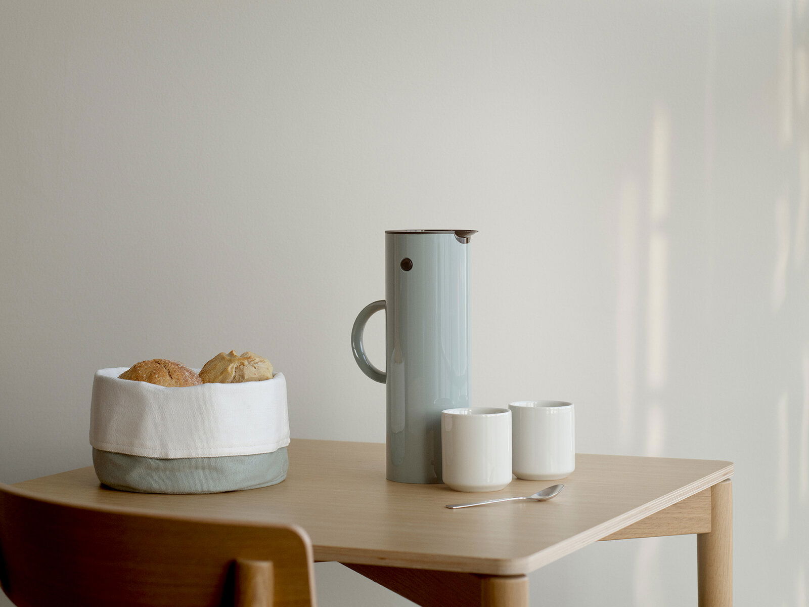 Stelton Isolierkanne 1 Liter Dusty Green Isolierkanne 1l in staubigem Grün auf einem Tisch, umgeben von zwei weißen Tassen, einem Löffel und einem Brotkorb. Seitliche Perspektive.