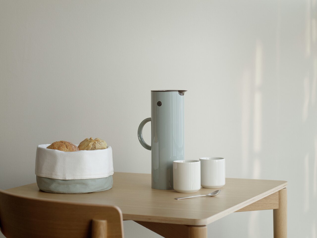 Stelton Isolierkanne 1 Liter Dusty Green Isolierkanne 1l in staubigem Grün auf einem Tisch, umgeben von zwei weißen Tassen, einem Löffel und einem Brotkorb. Seitliche Perspektive.