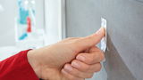 Nahaufnahme einer Hand, die einen tesa Powerstrip Waterproof an einer glatten, grauen Wand befestigt. Perspektive von der Seite.