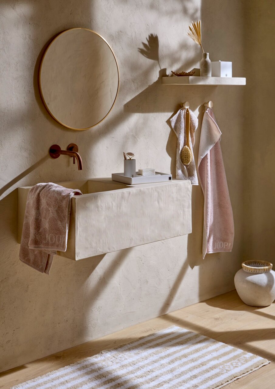 JOOP! Duschtuch DOUBLEFACE Badezimmer mit elegantem, beigem Waschbecken, runder Spiegel, zwei hängende Duschtücher in Rosa und Beige, dekorative Elemente auf einem Regal, seitliche Perspektive.