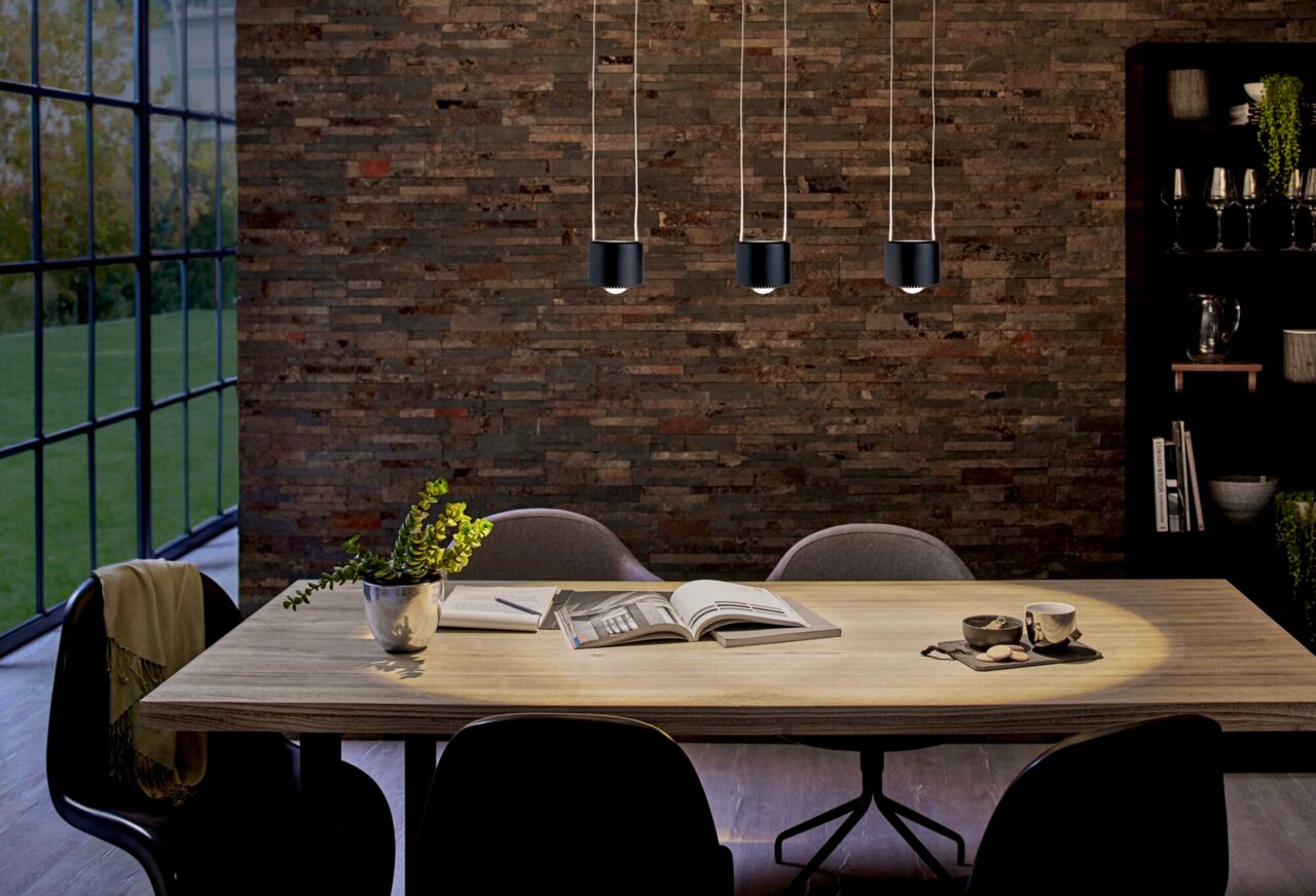 Drei moderne, schwarze Pendelleuchten über einem Esstisch in einem stilvollen Esszimmer mit Backsteinwand und großen Fenstern, seitliche Perspektive.