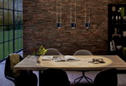 Drei moderne, schwarze Pendelleuchten über einem Esstisch in einem stilvollen Esszimmer mit Backsteinwand und großen Fenstern, seitliche Perspektive.